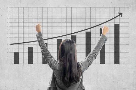 Rear view of Asian businesswoman looking at increased business finance graph with wall backgroundの写真素材