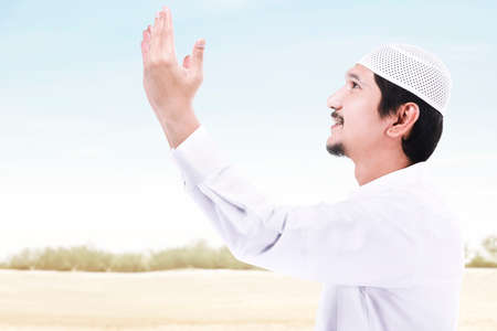 Asian Muslim man standing while raised hands and praying with a blue sky backgroundの写真素材