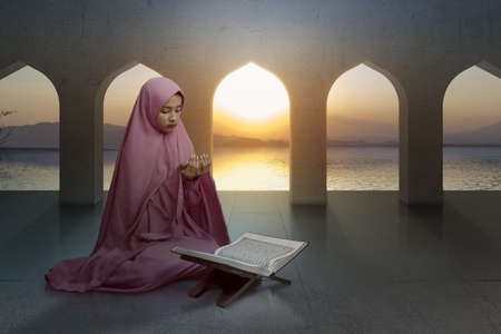 Asian Muslim woman in a veil sitting while raised hands and praying in the mosqueの写真素材
