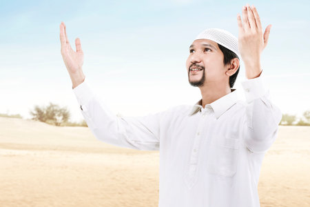 Asian Muslim man standing while raised hands and praying with a blue sky backgroundの写真素材