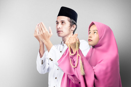 Asian Muslim couple standing while raised hands and praying together with gray wall background