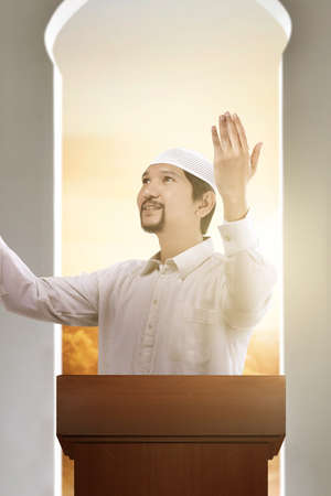 Asian Muslim man standing while raised hands and giving a sermon on the mosque