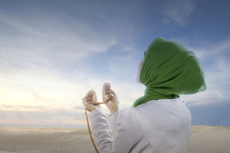 Asian Muslim woman in veil praying with prayer beads on her hands with a blue sky backgroundの写真素材
