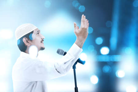 Asian Muslim man standing while raised hands and giving a sermon with blurred light background