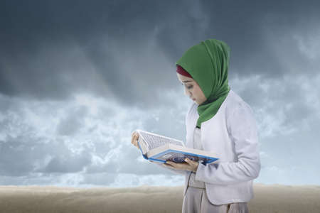 Asian Muslim woman in a veil standing and reading the Quran with a dramatic sky backgroundの写真素材