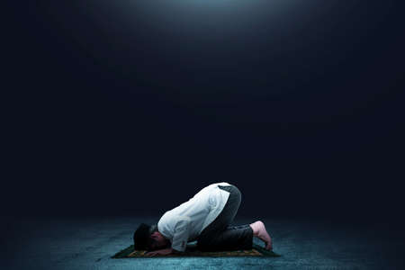 Asian Muslim man in praying position (salat) on prayer rug with dark background