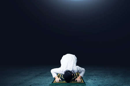 Asian Muslim man in praying position (salat) on prayer rug with dark background