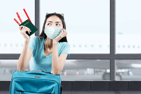 Asian woman in the face mask with a suitcase holding ticket and passport on the airport terminal. Traveling in the new normalの写真素材