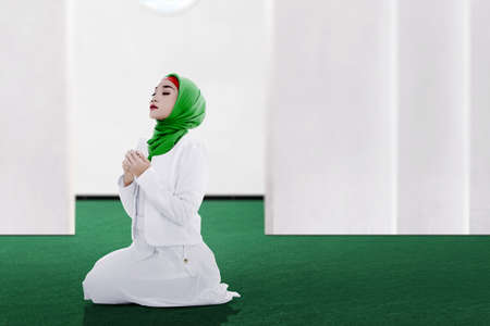 Asian Muslim woman in a veil sitting while raised hands and praying on the mosqueの写真素材