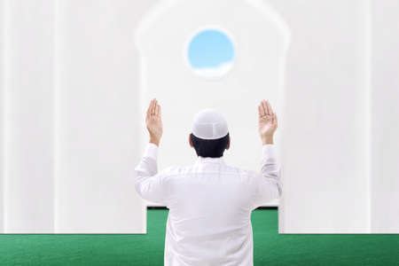 Rear view of Asian Muslim man sitting while raised hands and praying on the mosqueの写真素材