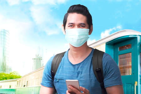 Asian man in the face mask with a backpack holding smartphone on the train station. Traveling in the new normalの写真素材