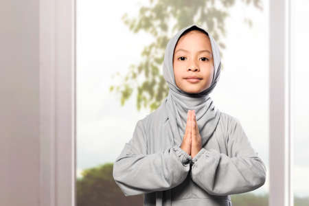 Asian Muslim girl in veil with greeting gesture at homeの写真素材