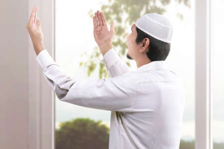 Rear view of Asian Muslim man sitting while raised hands and praying at homeの写真素材