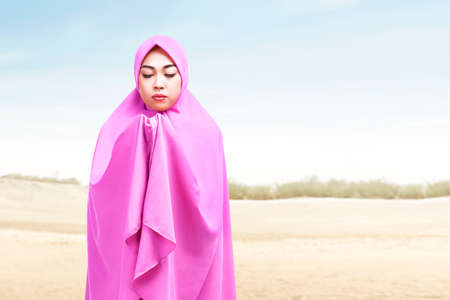 Asian Muslim woman in a veil standing while raised hands and praying on the duneの写真素材