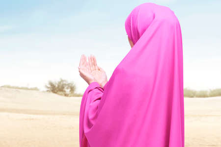 Rear view of Asian Muslim woman in a veil standing while raised hands and praying on the duneの写真素材