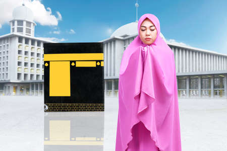 Asian Muslim woman in a veil standing and praying with Kaaba view and blue sky background