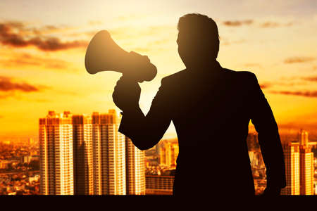 Silhouette of a businessman standing and using megaphone with cityscapes backgroundの写真素材