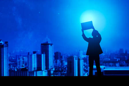 Silhouette of a businessman standing and holding a board with cityscapes backgroundの写真素材