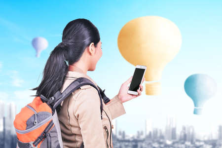 Asian woman with backpack and smartphone looking at colorful air balloon flying with cityscape backgroundの写真素材