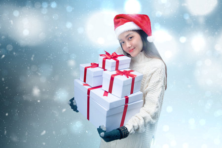 Asian woman with winter gloves and Santa hat holding white gift box with blurred light background