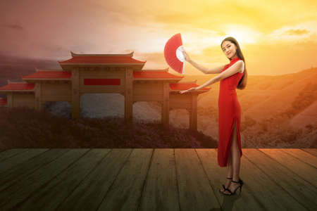 Asian Chinese woman in a cheongsam dress holding fan with open palm showing something with a dramatic sky background. Happy Chinese New Yearの写真素材