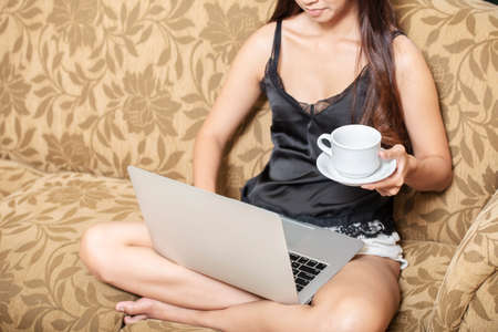 Asian woman sitting while using a laptop and holding a cup of coffee on the couchの写真素材