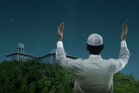 Rear view of Asian Muslim man standing while raised hands and praying on the nightの写真素材