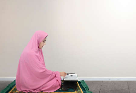 Asian Muslim woman in a veil sitting and reading the Quran with white wall backgroundの写真素材