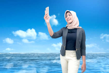 Asian Muslim woman in a headscarf taking a selfie with her mobile phone with blue sky backgroundの写真素材