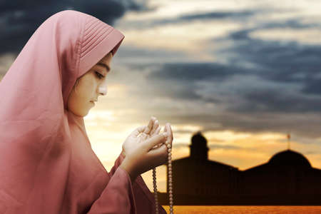 Asian Muslim woman in veil praying with prayer beads on her hands with the dramatic scene background