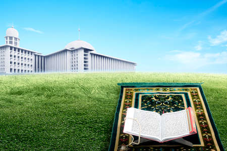 Quran open on a wooden placemat on a prayer rug on the field with a mosque view