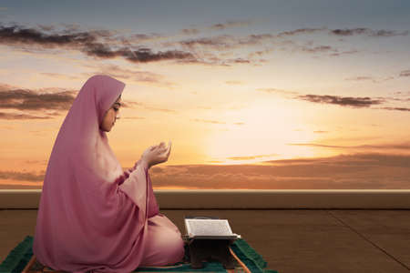 Asian Muslim woman in a veil sitting while raised hands and praying in the dramatic scene backgroundの写真素材
