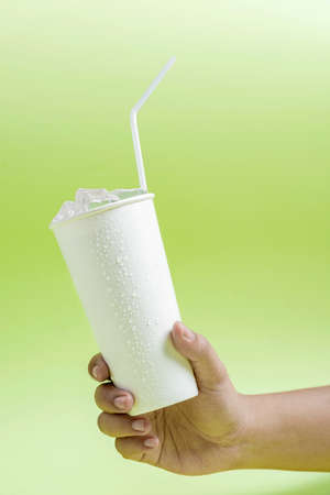 Human hand holding a white plastic cup with ice cubes and a straw with colored background