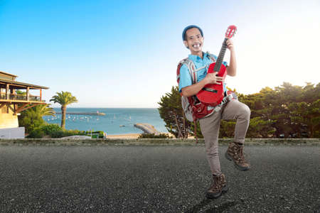 Asian man with a beanie hat and a backpack carrying a guitar standing on the street with a beach...