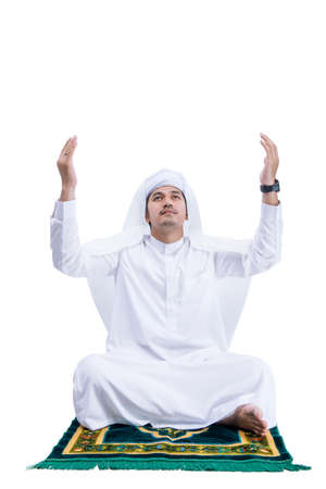 Muslim man praying while sitting on the prayer rug isolated over white backgroundの写真素材