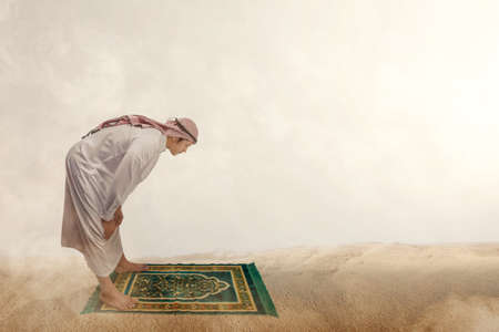 Muslim man with keffiyeh with agal in praying position (salat) on the prayer rug in the sand duneの写真素材