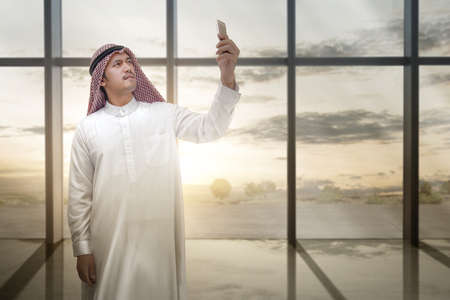 Arab man wearing keffiyeh looking at mobile phone inside the room with window glass and sunset sky...