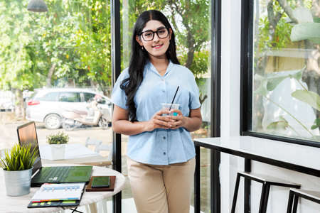 Asian woman in eyeglasses with a laptop and notebook on the table holding coffee while standing in...