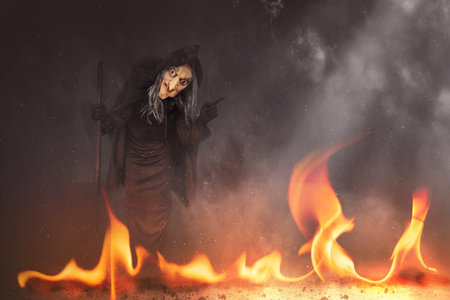 Old witch in a cloak with a stick standing with a fire background. Halloween concept