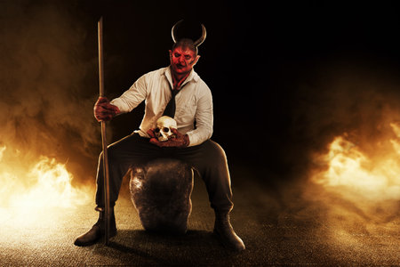 Devilman sitting while holding sticks and a human head skull with fire's background. Halloween...