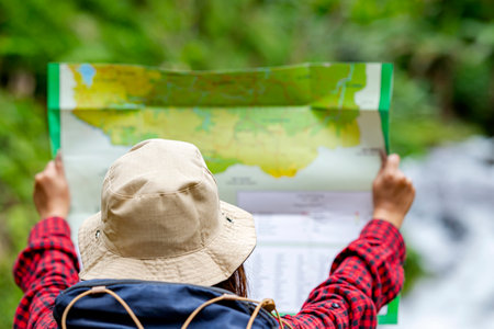 Rear view of Asian woman in a hat and backpack holding maps in the forest