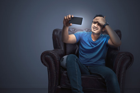 Asian man with the ball sitting on the couch with a disappointed expression watching a football match from a mobile phone at homeの写真素材