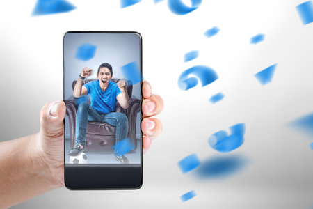 Asian man with the ball sitting on the couch with an excited expression watching football on the mobile phone screenの写真素材