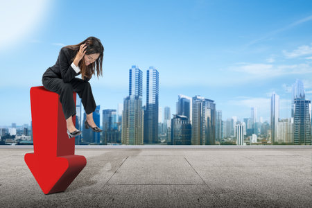 Asian businesswoman sitting on down arrow with frustrated expression with cityscape backgroundの写真素材