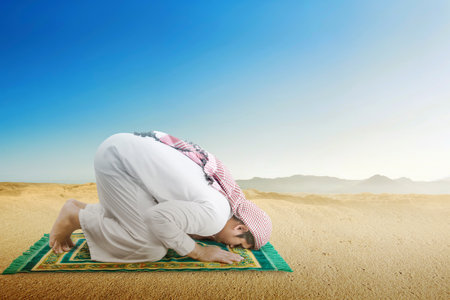 Muslim man with keffiyeh and agal in praying position (salat) on the prayer rug in the desertの写真素材