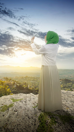 The rear view of a Muslim woman in a veil raising a hand and praying in the dramatic sky backgroundの写真素材