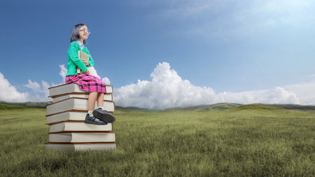 Asian little girl with eyeglasses holding a book and sitting on the stack of books. World Book Day Conceptの写真素材