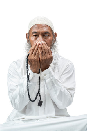 Muslim man with a beard wearing a white cap praying with prayer beads on his hands while sitting isolated over white backgroundの写真素材