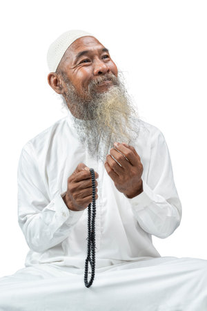 Muslim man with a beard wearing a white cap praying with prayer beads on his hands while sitting...