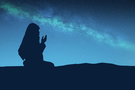 Silhouette of a Muslim man with a beard wearing keffiyeh with agal in praying while raised hands with the night scene backgroundの写真素材